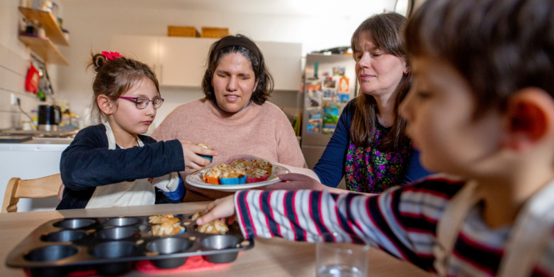 Eine blinde Mutter sitzt mit drei weiteren Personen an einem Tisch, sie bereiten Muffins vor.