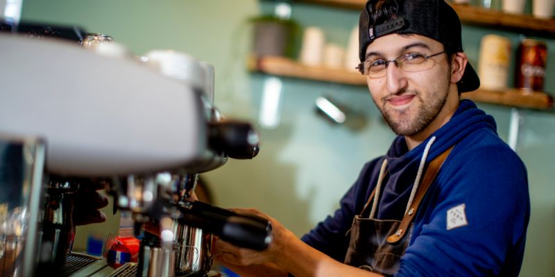 Ein junger Mann mit Baseball-Kappe bedient eine Kaffeemaschine in einem Lokal.