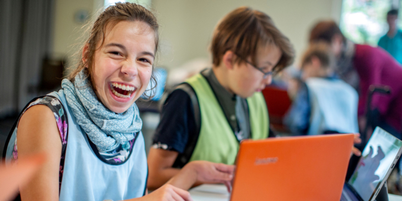 Zwei Kinder mit Behinderung sitzen an ihren Laptops. Das Mädchen lächelt, der Junge arbeitet am Bildschirm.
