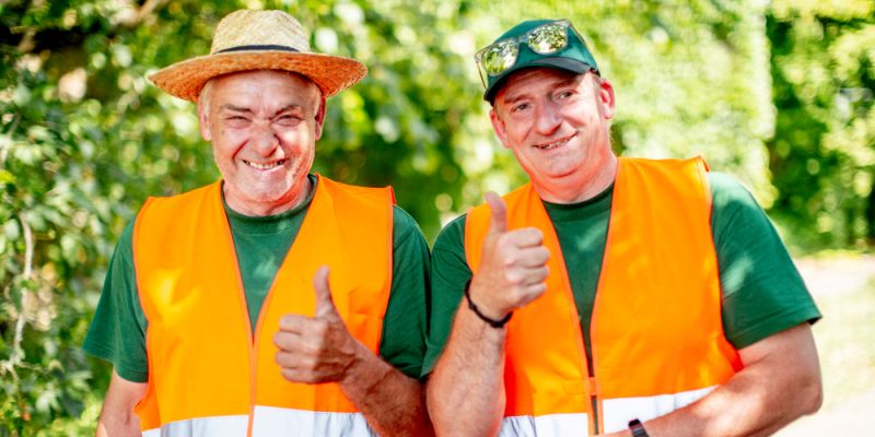 Zwei Gartenarbeiter in orangefarbenen Warnwesten lachen in die Kamera und zeigen den Daumen hoch.