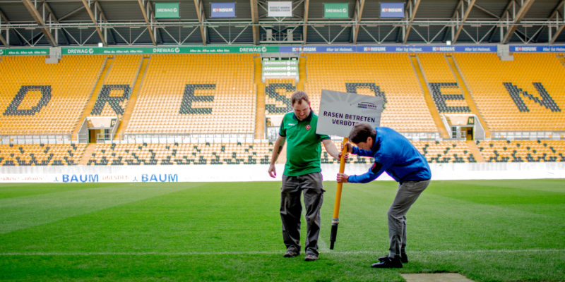 Zwei Männer stecken ein Schild mit der Aufschrift "Betreten verboten" in den Rasen eines Fußballfeldes.