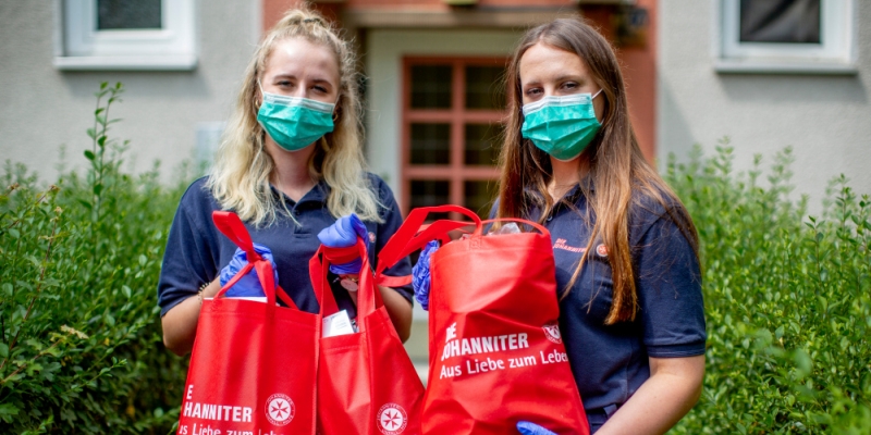 Zwei junge Frauen mit Mundschutz halten mit Einkäufen gefüllte Stoffbeutel der Johanniter in die Höhe.