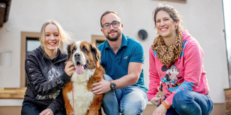 Zwei Frauen und ein Mann lächeln in die Kamera und streicheln einen großen Hund.