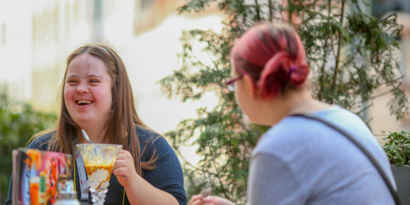 Zwei Mädchen mit und ohne Behinderung sitzen an einem Tisch in einem Caf, essen Eis und lachen.
