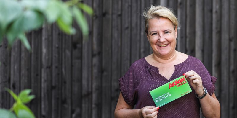 Eine Frau hält ein Aktion Mensch-Los in Händen und lacht in die Kamera.