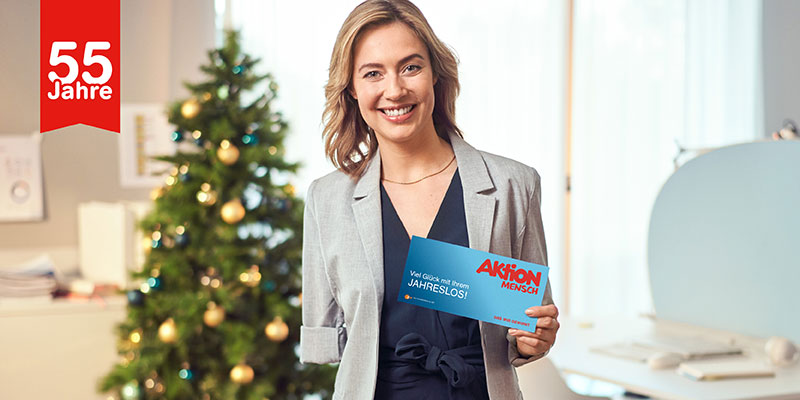 Eine Frau mit einem Arm steht vor einem Weihnachtsbaum und hält ein Los der Aktion Mensch in einer Hand.