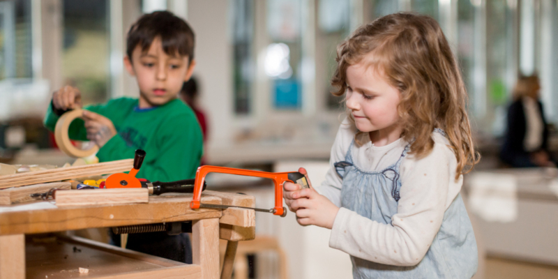 Zwei Kinder stehen an einer Werkbank. Ein Mädchen hat eine kleine Säge in der Hand.