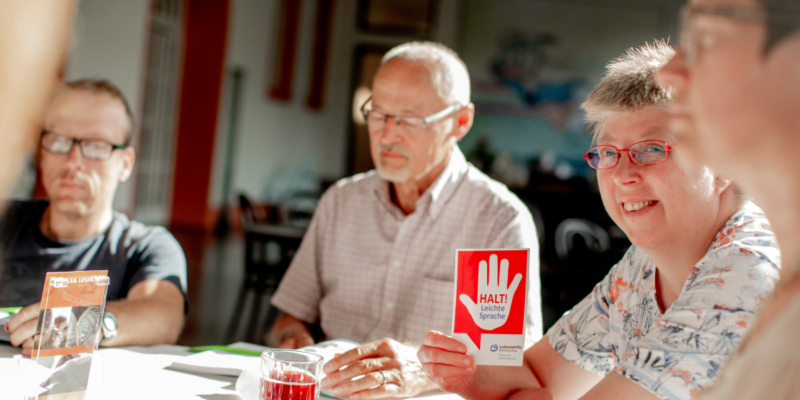 Mehrere Menschen sitzen an einem Tisch in einer Leserunde. Eine Frau hält eine Postkarte mit der Aufschrift "Halt! Leichte Sprache" in den Händen.