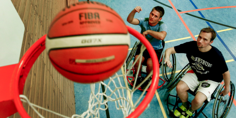 Zwei junge Männer in Rollstühlen stehen unter einem Basketballkorb und haben gerade einen Treffer gezielt.