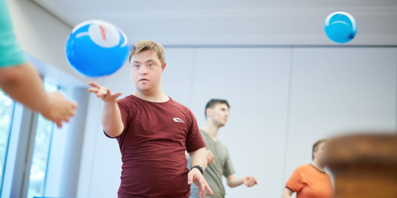 Ein junger Erwachsener wirft einen Ball in einer Turnhalle in die Luft.
