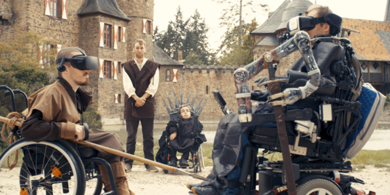 Eine mittelalterliche Kulisse: Zwei junge Männer in Rollstühlen duellieren sich mit Virtual Reality-Brillen. Im Hintergrund schauen zwei Männer mit und ohne Behinderung zu.