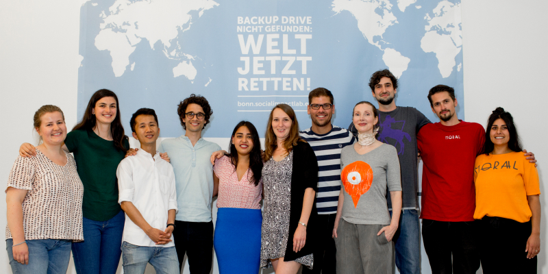 Junge Männer und Frauen stehen vor einem großen Banner mit der Aufschrift "Backup Drive nicht gefunden: Welt jetzt eetten!".