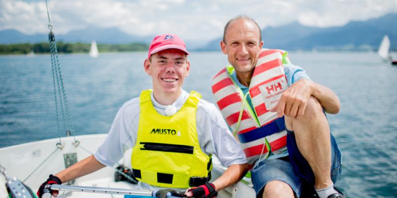 Ein Jugendlicher mit Behinderung und ein Mann sitzen auf einem Segelboot und lächeln in die Kamera.