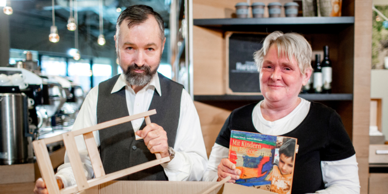 Ein Mann und eine Frau stehen vor einer Theke und lächeln in die Kamera. Der Mann hält ein kleines Holzgestell in den Händen, die Frau ein Buch mit der Aufschrift "Mit Kindern im Bienengarten".
