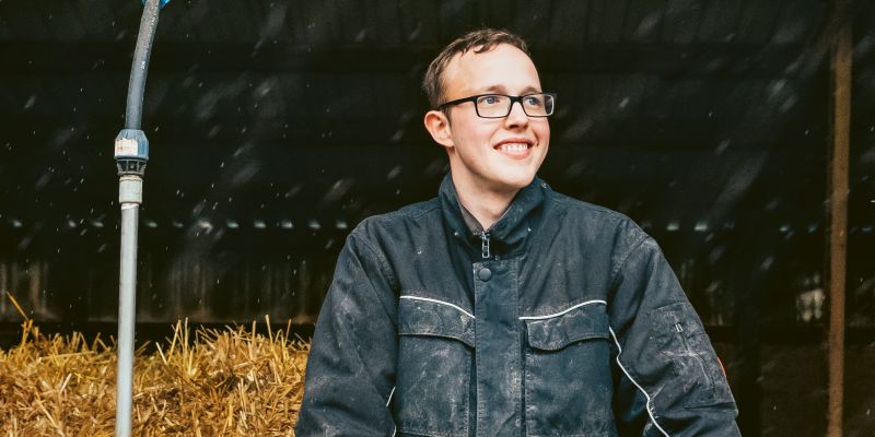 Ein junger Mann mit Brille und Arbeitskleidung steht in einer Scheune.