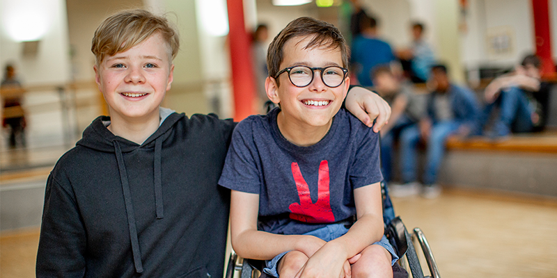 Zwei Jungen im Teenageralter, einer mit und einer ohne Rollstuhl, sitzen nebeneinander und halten sich lachend im Arm.