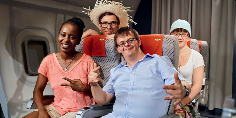 Shary Reeves sitzt in einem Flugzeug. Neben ihr sitzen ein Mann und eine Frau mit Down-Syndrom. Sie lächeln in die Kamera. In der zweiten Reihe sitzt ein Mann mit Strohhut.