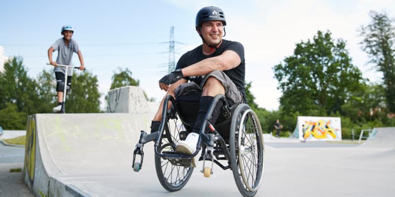 Ein junger Mann sitzt in Skater-Montur im Rollstuhl auf einer Skate-Anlage und lächelt in die Kamera