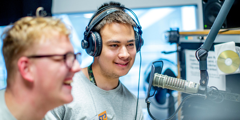 Zwei junge Männer stehen in einem Radiostudio mit Koprfhörern auf den Ohren vor einem Mikrofon und Sprechen Text ein.