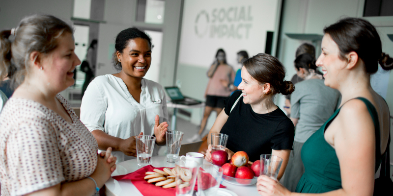 Vier Frauen stehen an einem Tisch und unterhalten sich. Auf einer Leinwand im Hintergrund ist der Schriftzug "Social Impact" zu sehen.