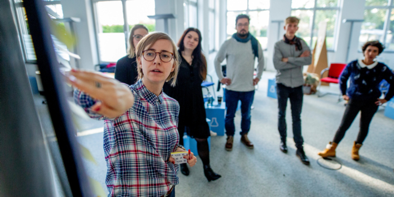 Eine Frau steht seitlich vor einem Whiteboard und erklärt etwas. Im Hintergrund sind fünf Personen zu sehen, die ihr zuhören.
