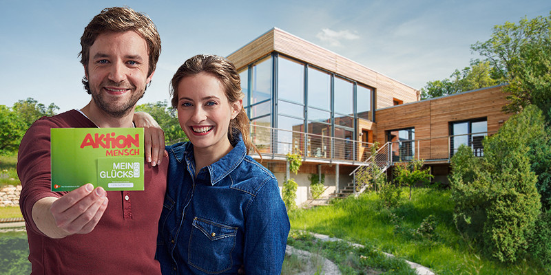 Ein Paar steht vor einem Traumhaus und hält ein Glückslos in den Händen