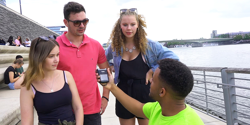 Ein junger YouTube-Journalist im Rollstuhl hält einer Gruppe von zwei Mädchen und einem jungen an den Rheintreppen in Köln ein Mikrofon hin, sie reden miteinnander.