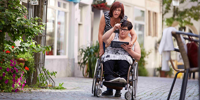 Frau schiebt eine Frau im Rollstuhl, die ein Tablet in der and hält