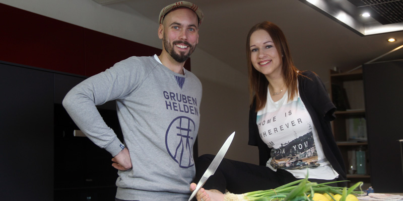 Ein Mann mit Mütze und eine Frau an einer Küchentheke. Die Frau sitzt auf der Theke und hält ein Küchenmesser mit den Zehen. Sie hat ein Bein und keine Arme.