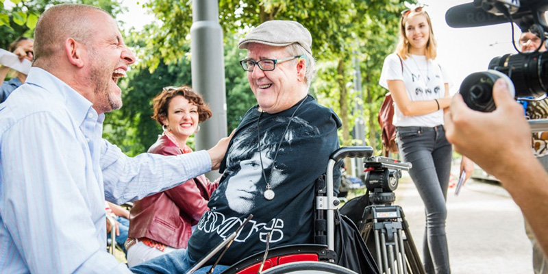 Zwei Männer lachen gemeinsam, einer der beiden sitzt im Rollstuhl. Die beiden werden von einer Kamera gefilmt.