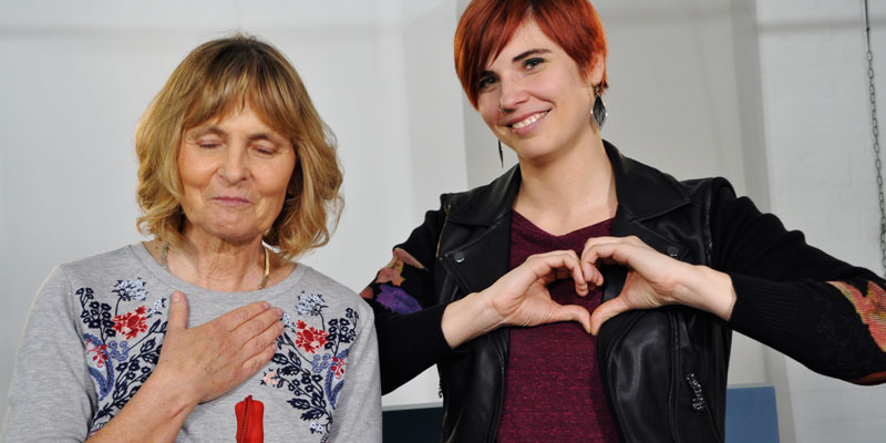 Eine blinde Frau und eine junge Frau. Die junge Frau macht ein Herz mit ihren Händen.