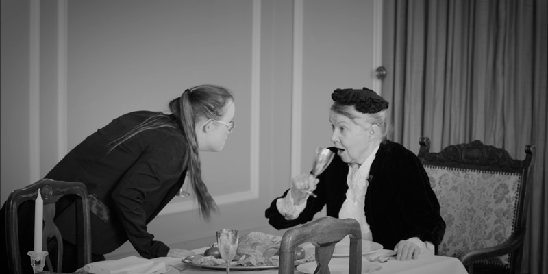 Eine Frau mit Down-Syndrom schaut einer älteren Frau, die aus einem Weinglas trinkt, in die Augen. Die ältere Frau sitzt am Tisch.