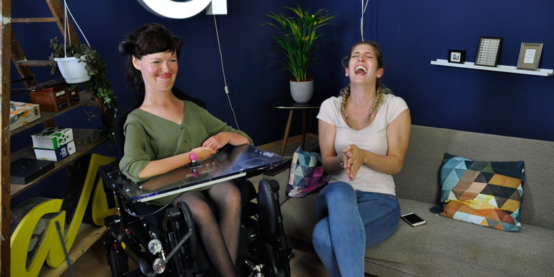 Eine Frau im Rollstuhl und eine blonde Frau lachen gemeinsam.