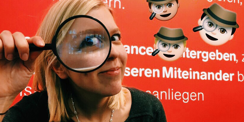 Eine blonde Frau sieht durch eine Lupe in die Kamera. Neben ihr sieht man Emojis mit Hut und Lupe.
