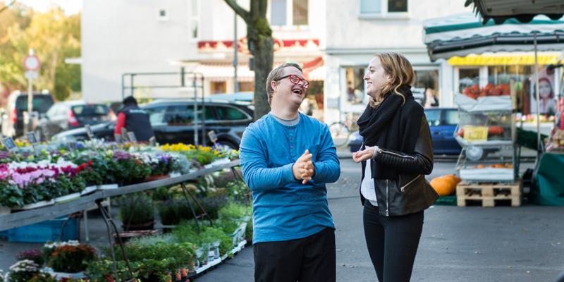Ein Mann mit Behinderung und eine Frau lachen auf dem Wochenmarkt.