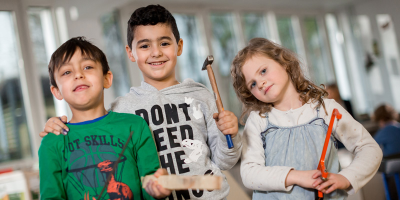 Drei Kinder lächeln in die Kamera. Sie halten Werkzeuge in der Hand.