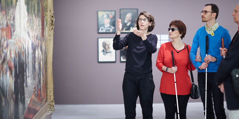 Eine Frau steht vor einem Bild im Museum und erzählt. Neben ihr hören Menschen mit Blindenstock zu.