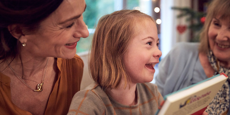 Eine Frau, eine ältere Frau und ein Mädchen mit Down-Syndrom lachen und sitzen auf dem Sofa. Das Mädchen hält ein Buch in der Hand.