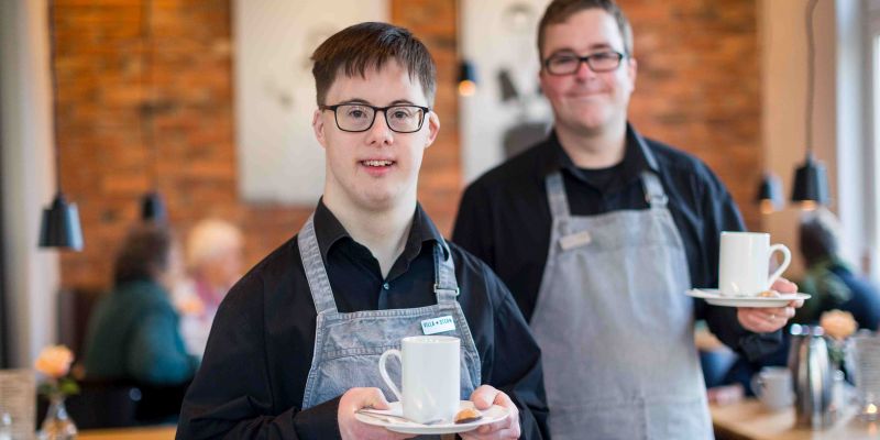 Zwei junge Männer mit Behinderung stehen in einem Frühstücksraum eines Hotels.