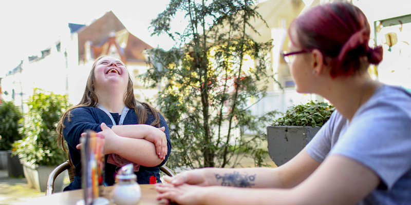 Zwei Frauen sitzen in einer Eisdiele und lachen gemeinsam. Eine der Frauen hat eine geistige Behinderung.