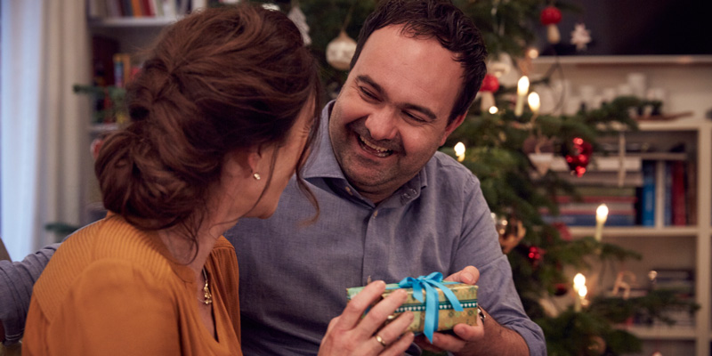 Eine Frau und ein Mann sitzen am Weihnachtsbaum. Die Frau überreicht dem Mann ein Geschenk.