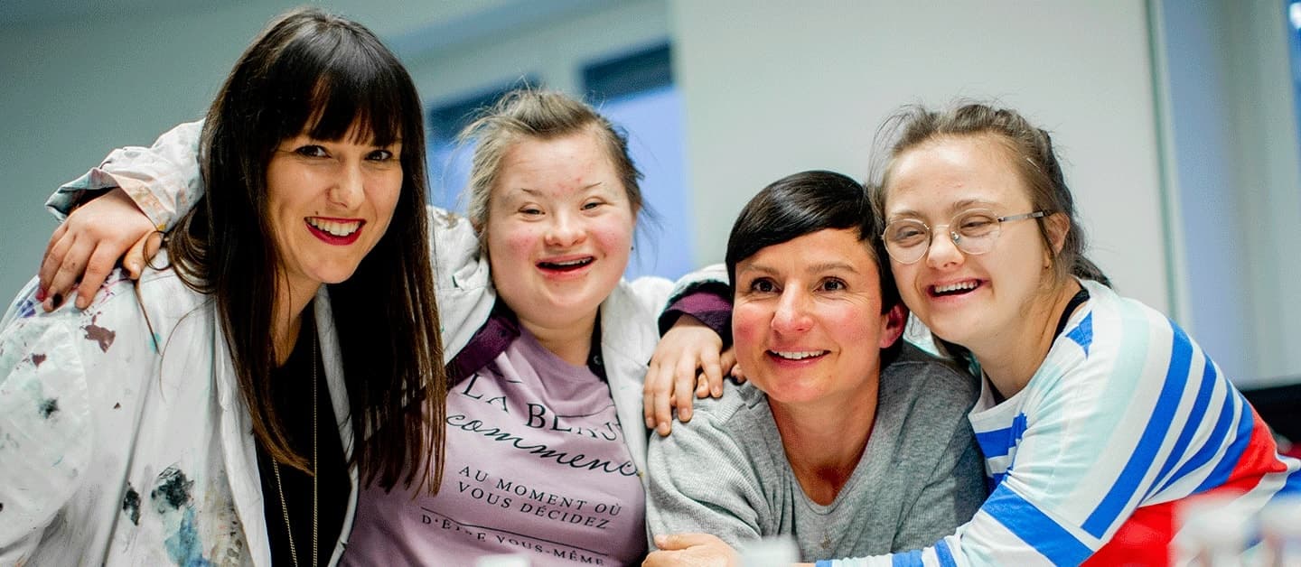 Vier Personen stehen eng beieinander, zwei mit Armen um die anderen, alle lächelnd.