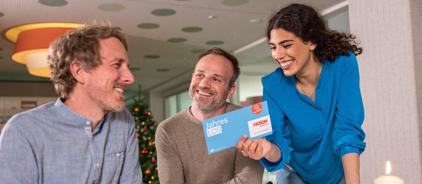 Drei Personen freuen sich über ein Aktion Mensch-Jahreslos vor einem Weihnachtsbaum.