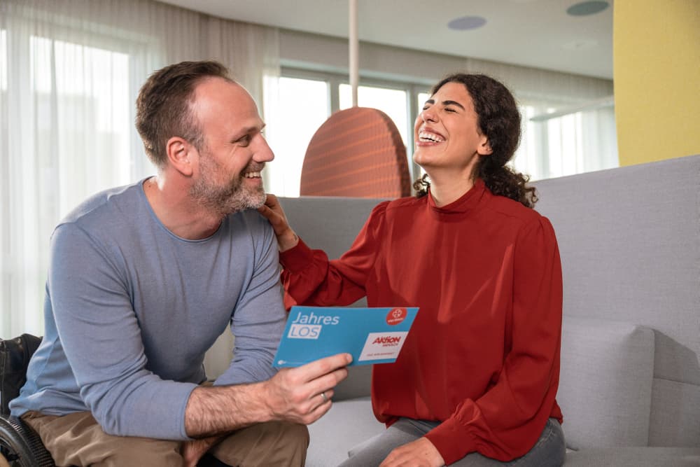 Ein Mann im Rollstuhl und eine Frau auf dem Sofa lachen sich herzlich an. Die beiden Mitarbeiter freuen sich über ihr Aktion Mensch-Los.
