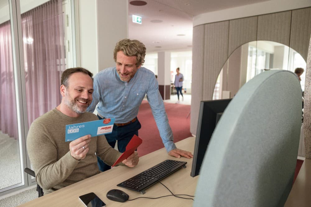 Ein Vorgesetzter schaut zu, wie ein Mitarbeiter sein Los von Aktion Mensch öffnet. Der Mitarbeiter freut sich über sein Los.