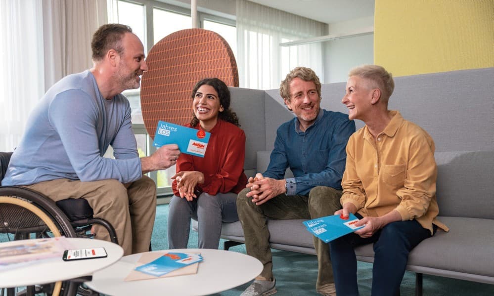 Vier Kollegen sitzen in einem Büro und unterhalten sich gut gelaunt. Eine Person sitzt im Rollstuhl. Sie halten Jahreslose von Aktion Mensch in der Hand.