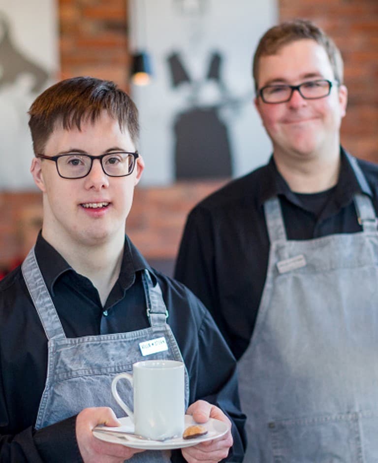 Zwei Männer in Schürzen stehen zusammen. Der Mann im Vordergrund hält eine Tasse Kaffee in der Hand.