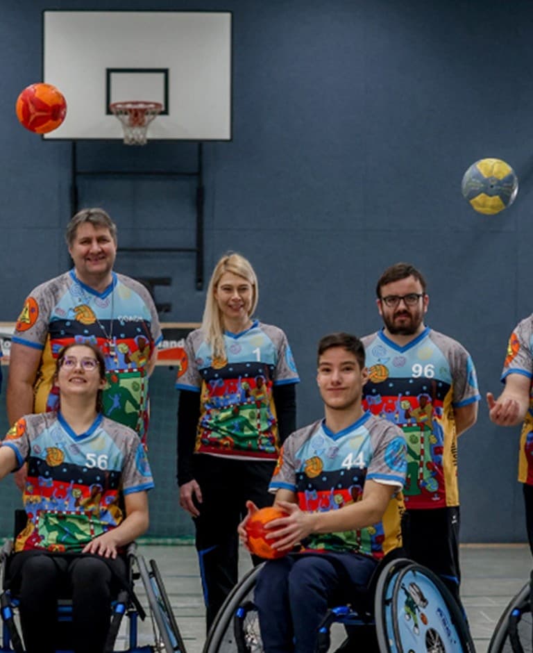 Eine Gruppe von Menschen halten Handbälle in der Hand und werfen sie in die Luft. Die Personen im Vordergrund sitzen im Rollstuhl.