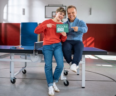 Niko Kappel und eine junge Frau sitzen entspannt auf einer Tischtennisplatte in einer Sporthalle und lächeln in die Kamera. Die Frau hält das grüne Glücks-Los der Aktion Mensch in der Hand, mit der anderen Hand zeigt sie darauf. Niko zeigt ebenfalls mit einer Hand auf das Los.