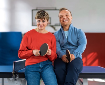 Niko Kappel und eine junge Frau sitzen entspannt auf einer Tischtennisplatte in einer Sporthalle. Beide halten Tischtennisschläger in der Hand und lächeln in die Kamera. Die Person links trägt einen roten Pullover und Jeans, Niko Kappel trägt eine blaue Sportjacke.